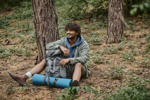 carefree young indian tourist resting while sitting near backpack on ground in forest