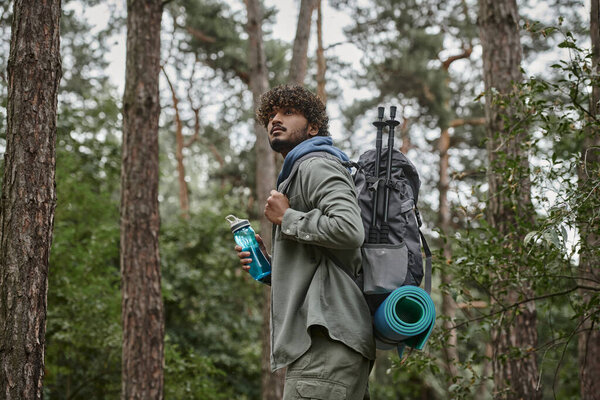 young curly indian tourist holding backpack and sports bottle in forest