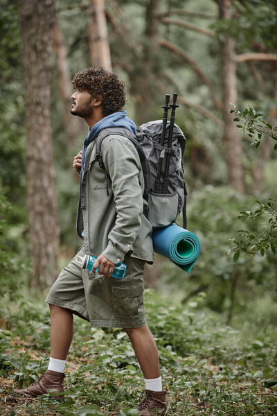 side view of young indian tourist with backpack and sports bottle walking in forest