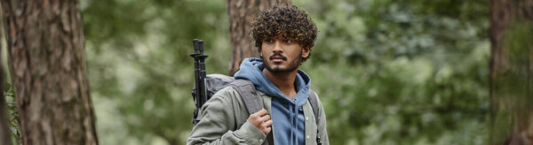 young curly indian tourist with backpack and trekking poles looking away in forest, banner