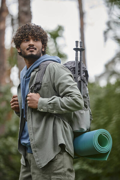young indian tourist with backpack and hiking supplies looking away in blurred forest
