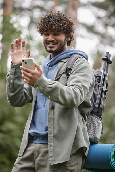 overjoyed indian hiker with backpack waving hand while having video call on smartphone in forest