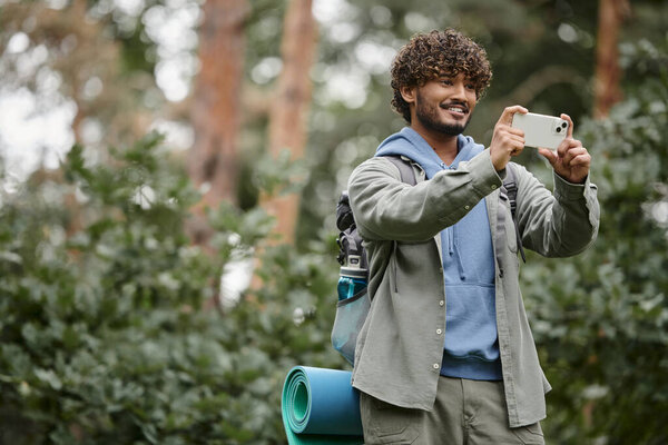 cheerful young indian hiker with backpack taking photo on smartphone in blurred forest