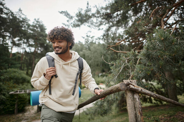 smiling young indian hiker with backpack walking near fence in blurred forest