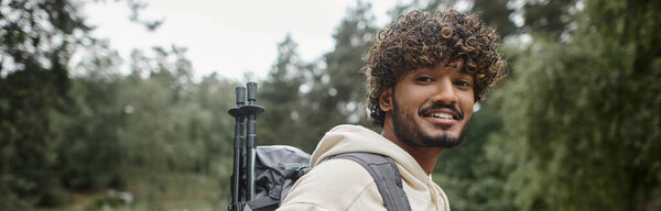 positive young indian traveler with trekking poles and backpack standing in forest, banner