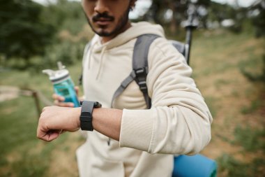 cropped view of indian tourist with backpack holding sports bottle and using smartwatch outdoors clipart