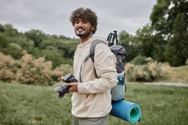 Ormanda, fotoğrafçılıkta ve macera konseptinde gülümseyen ve dijital kamera tutan Hintli sırt çantalı.