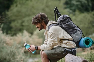 Sırt çantasıyla oturan mutlu Hintli turist ormanda suyla dolu bir şişe tutuyor.