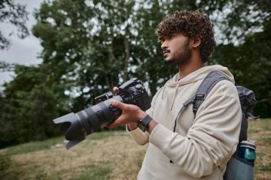 concentrated indian man holding digital camera and standing with backpack outdoors, photographer clipart