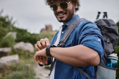happy indian man in sunglasses and denim shirt checking time on wristwatch, hiker with travel gear clipart