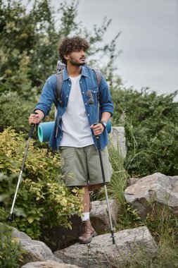 curly indian tourist walking with backpack and holding hiking sticks during trekking, wild nature clipart