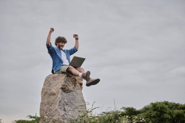 Dijital göçebe konsepti, heyecan verici Hintli serbest yazar kazananı kutlarken dizüstü bilgisayar kullanıyor, kaya üzerinde oturuyor