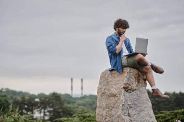 Dijital göçebe, kıvırcık Hintli serbest bilgisayar kullanan, kaya üzerinde oturan, uzaktan çalışma konsepti