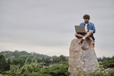 Dijital göçebe, kıvırcık Hintli adam dizüstü bilgisayar kullanıyor, kayanın üzerinde oturuyor, uzaktan çalışma kavramı, manzara görüntüsü