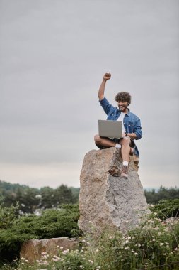 Dijital göçebe kavramı, heyecanlı Hintli adam kazanmayı kutlarken dizüstü bilgisayar kullanıyor, kayanın üzerinde oturuyor.