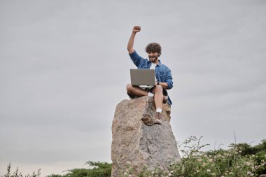 digital nomad concept, amazed indian freelancer covering mouth and using laptop, sitting on rock clipart