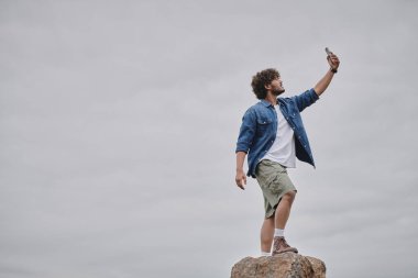 Nomadizm konsepti, kıvırcık Hintli adam kayanın üzerinde duruyor ve akıllı telefonu tutarken sinyal arıyor.