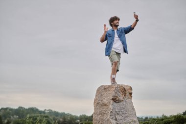 Nomadizm konsepti, mutlu Hintli adam kaya üzerinde duruyor ve akıllı telefonda görüntülü sohbet ediyor, el sallıyor.