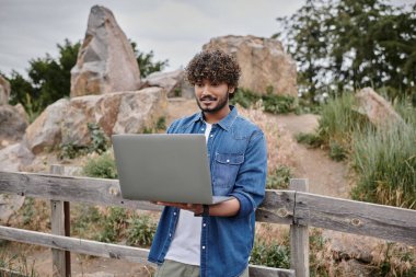 Dijital göçebe kavramı, mutlu Hintli adam tahta çitin yanında duruyor ve dizüstü bilgisayar kullanıyor, kırsal kesimde.