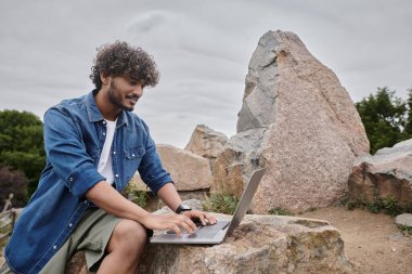 happy indian man typing on laptop and working remotely in natural place, digital nomadism clipart