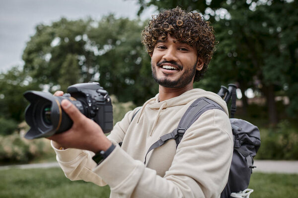 happy indian photographer with backpack taking photo on professional camera, adventure and travel