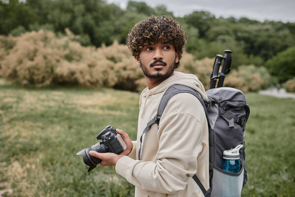 curly indian photographer with backpack holding professional camera outdoors, adventure and travel
