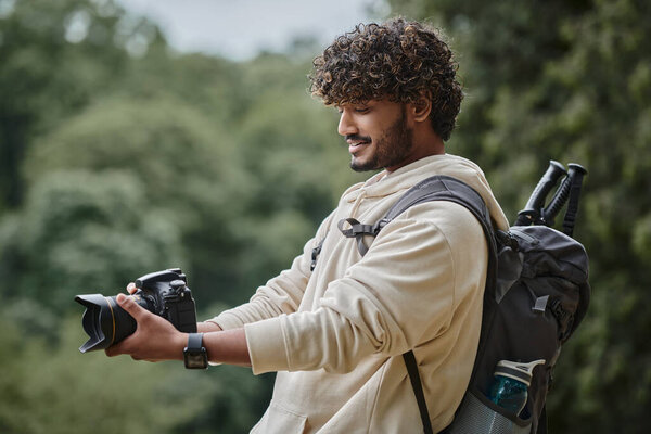 joyful indian tourist taking photo on professional camera, tourist with backpack in forest