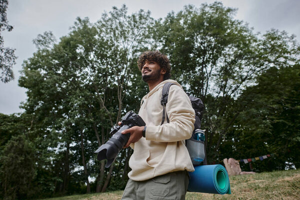 happy indian man holding digital camera and standing with backpack in forest, photographer