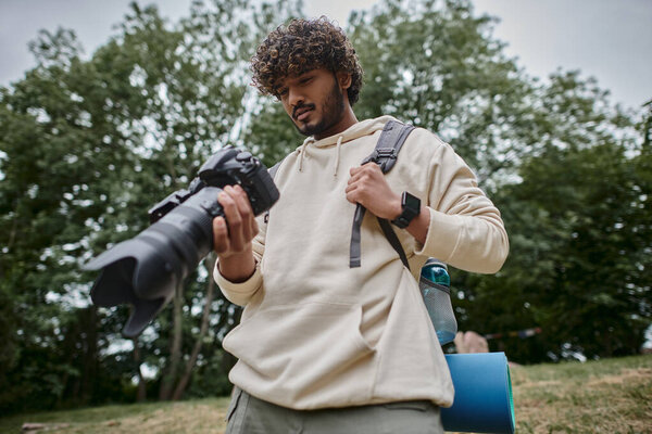 positive indian man holding digital camera and standing with backpack in forest, photographer