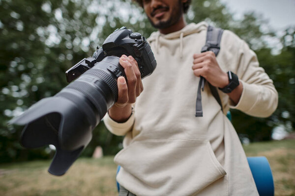 cropped view of indian man holding digital camera and standing with backpack in woods, photographer