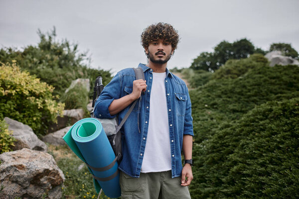 curly indian man standing with backpack and sleeping mat during trip, hiker with travel gear