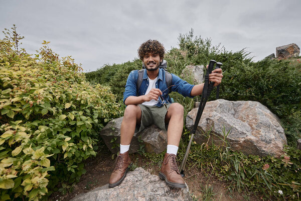 indian man sitting with backpack on rock and holding hiking sticks during trekking, adventure
