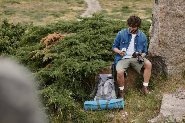 tourist and nature concept, indian man holding bottle with water and camera while sitting on rock