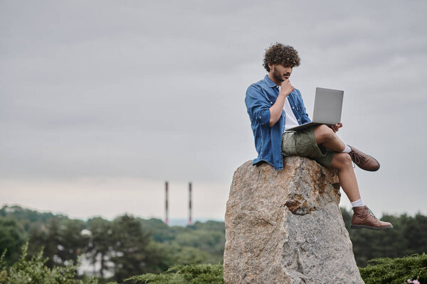 digital nomad, curly indian freelancer using laptop, sitting on rock, remote work concept