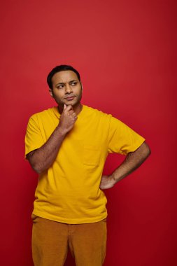 pensive indian man in bright casual attire standing with hand near chin on red background in studio clipart