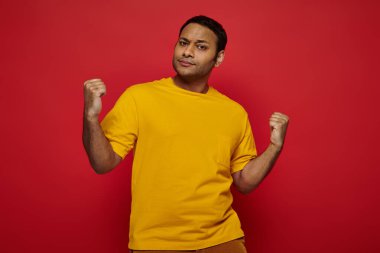 expressive indian man in casual clothes standing with clenched fists  on red background, strength clipart