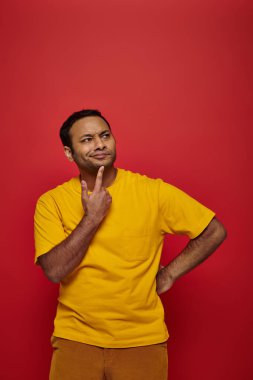 thoughtful indian man in bright casual clothes looking away on red background, creative idea clipart