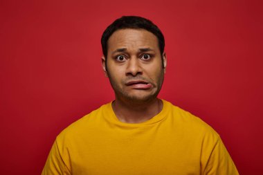 worried indian man in bright clothes looking at camera and grimacing on red background, stroke clipart