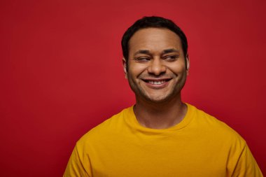 sly indian man in bright yellow clothes looking away and smiling on red background, side glance clipart