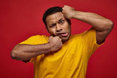 face expression, confused indian man in yellow t-shirt punching himself into face on red backdrop clipart