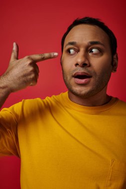 shocked indian man in yellow t-shirt pointing with finger at head on red background in studio clipart