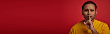 indian man in yellow t-shirt showing shh gesture on red backdrop, secret, finger near lips, banner clipart
