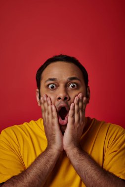 astonished indian man in yellow t-shirt looking at camera with wide open eyes on red backdrop clipart