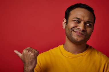 skeptical indian man in yellow t-shirt pointing with thumb and looking away on red backdrop clipart