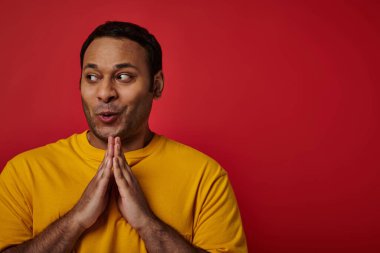 cunning indian man in yellow t-shirt clasping hands and planning something on red backdrop, sly face clipart