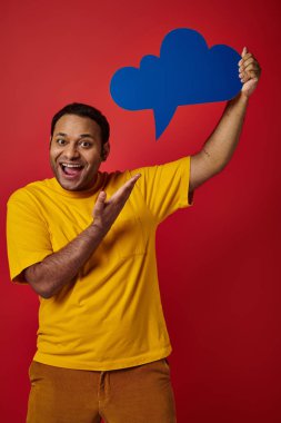 excited indian man in yellow t-shirt pointing blank thought bubble on red backdrop, happy face clipart