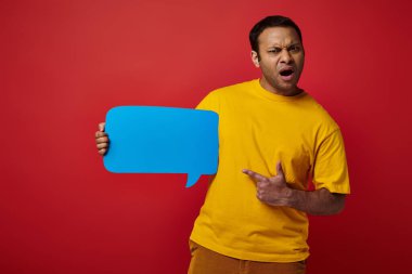 shocked man in yellow t-shirt pointing at blank speech bubble on red background, displeased clipart