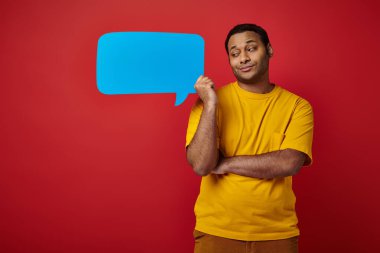 skeptical indian man in yellow t-shirt holding blank speech bubble on red background clipart