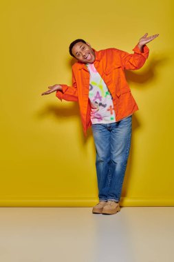 full length of positive indian man in orange jacket and denim jeans gesturing on yellow backdrop clipart
