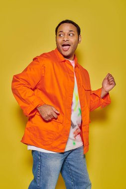portrait of amazed indian man in orange jacket running away and looking back on yellow backdrop clipart